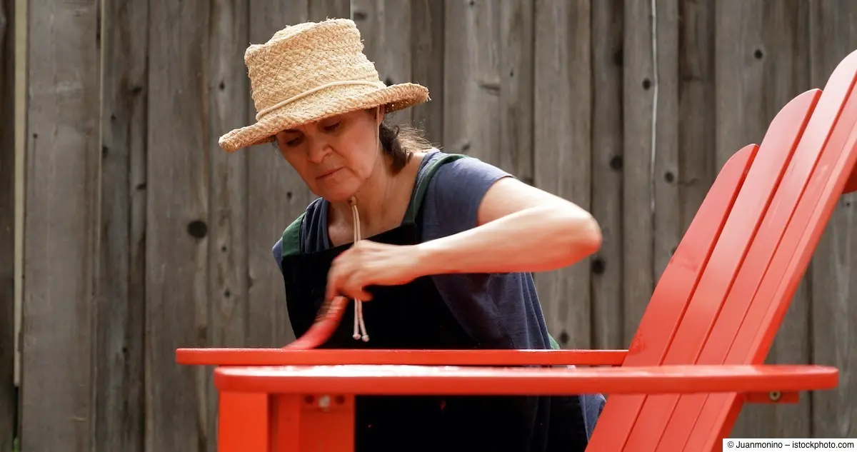 Frau mit Sonnenhut lackiert einen Gartenstuhl knallrot
