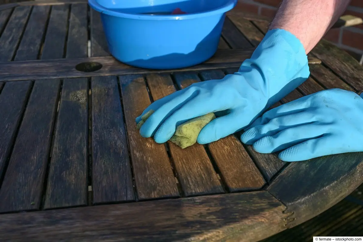 Person mit Handschuhen schrubbt verwitterten Holzgartentisch sauber Reinigung von verwitterten Holzgartenmöbeln mit Schwamm und Wasser, Person trägt blaue Gummihandschuhe für den Schutz.