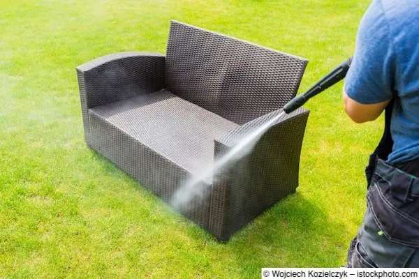 Mann säubert mit Hochdruckreiniger Gartensofa aus Polyrattan auf Wiese