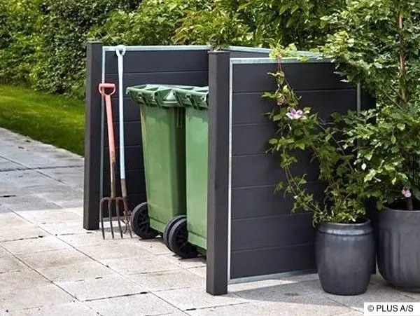 Dekorativer Sichtschutz für zwei Mülltonnen aus WPC-Elementen Moderne Mülltonnenverkleidung aus dunklen Paneelen mit Metallrahmen, umgeben von Pflanzen für eine ästhetische Gartengestaltung.