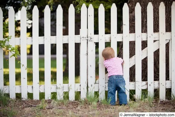Kleinkind steht an Gartenzaun und schaut durch die Latten